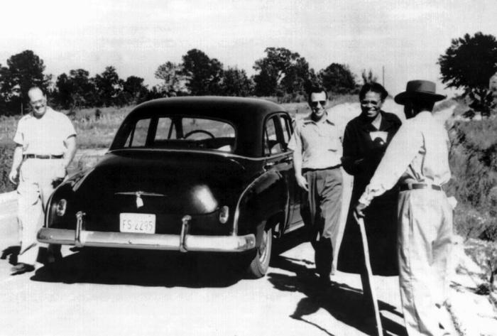 Four people beside a car on a rural road during a visit connected to the Tuskegee study; one man stands with his back to the camera holding a walking stick.